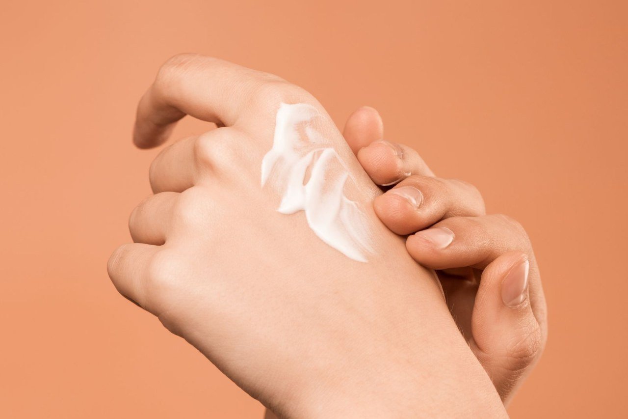 close-up of hands rubbing moisturizer cream into skin against a peach background
